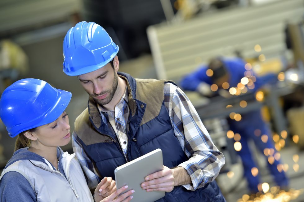 Two workers in blue hard hats review a tablet in a workshop while metal grinding continues behind.